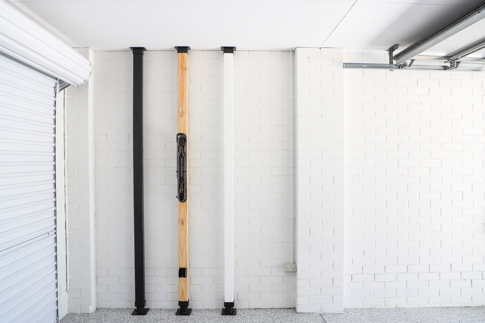 A set of three wooden posts with black brackets at the top and bottom, the middle one with a bike rack attached, with a white brick wall background.