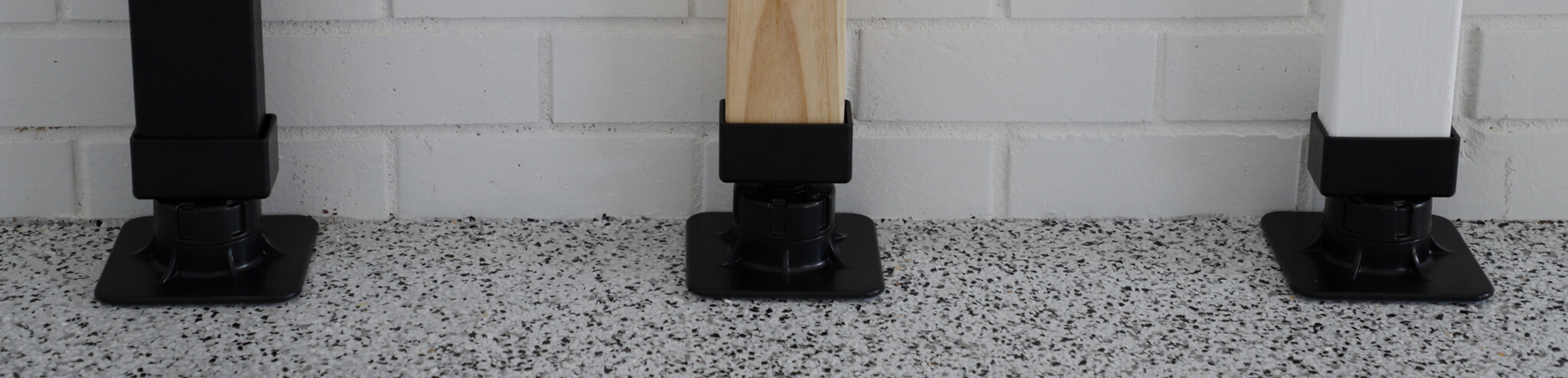 Three wooden posts with black bases on a speckled floor against a white brick wall.